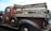 A vintage brown pickup truck with a wooden plank bed, showcasing old-fashioned design. The truck features a weathered look with visible rust and text on the wood panels. It has round, black tires with red rims and a classic 'Esso' logo on the driver's door.