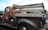 A vintage brown pickup truck with a wooden plank bed, showcasing old-fashioned design. The truck features a weathered look with visible rust and text on the wood panels. It has round, black tires with red rims and a classic 'Esso' logo on the driver's door.