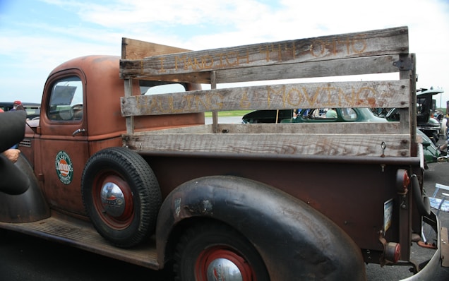 A vintage brown pickup truck with a wooden plank bed, showcasing old-fashioned design. The truck features a weathered look with visible rust and text on the wood panels. It has round, black tires with red rims and a classic 'Esso' logo on the driver's door.