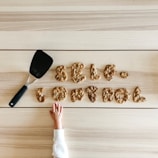 Cookies arranged to spell 'self control' rest on a wooden surface. A spatula is placed nearby, and a child's hand reaches towards the cookies.