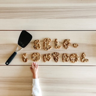 Cookies arranged to spell 'self control' rest on a wooden surface. A spatula is placed nearby, and a child's hand reaches towards the cookies.