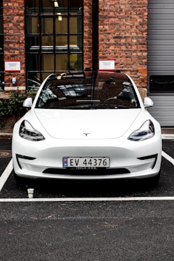 A white Tesla car is parked in a parking space in front of a brick building with large windows. The building has a mix of red and brown bricks and some visible signage. There is a small white cup on the ground in front of the car. The car has a Norwegian license plate.