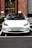 A white Tesla car is parked in a parking space in front of a brick building with large windows. The building has a mix of red and brown bricks and some visible signage. There is a small white cup on the ground in front of the car. The car has a Norwegian license plate.