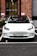 A white Tesla car is parked in a parking space in front of a brick building with large windows. The building has a mix of red and brown bricks and some visible signage. There is a small white cup on the ground in front of the car. The car has a Norwegian license plate.