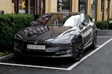 A sleek, glossy black Tesla car is parked in a designated spot outside a building with large windows and yellow walls. The vehicle's reflection is visible on the highly polished surface, and greenery lines the side of the parking area.