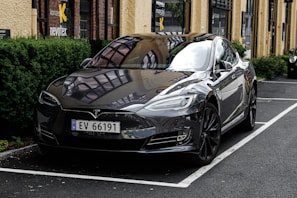 A sleek, glossy black Tesla car is parked in a designated spot outside a building with large windows and yellow walls. The vehicle's reflection is visible on the highly polished surface, and greenery lines the side of the parking area.