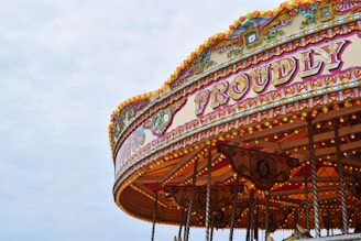 A vintage carousel decorated with elaborate patterns and bright lights is partially visible against a cloudy sky. The word 'PROUDLY' is prominently displayed in ornate lettering, surrounded by intricate designs and a warm, inviting glow from numerous bulbs.