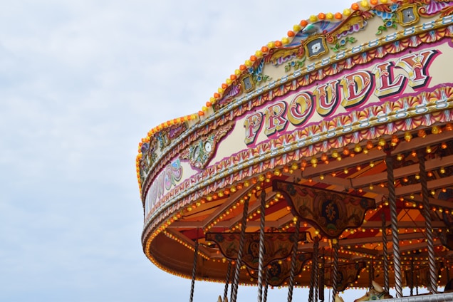 A vintage carousel decorated with elaborate patterns and bright lights is partially visible against a cloudy sky. The word 'PROUDLY' is prominently displayed in ornate lettering, surrounded by intricate designs and a warm, inviting glow from numerous bulbs.