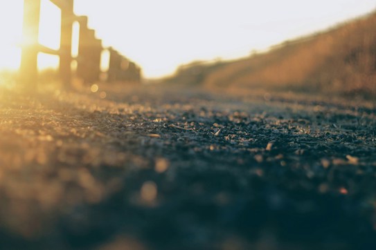 An out-of-focus image highlighting a sunlit pathway with a fence on the left. The warm glow of the sunlight creates a soft and serene ambiance, while the path leads off into the distance, suggesting a peaceful journey.