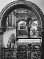 A black and white architectural scene featuring ornate arches and intricate decorative ironwork. The composition is symmetrical, showcasing multiple levels of arches with detailed carvings and patterns. Light filters through the arches, casting shadows and highlighting the elaborate designs.