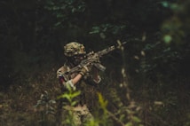 A person dressed in camouflage military attire, including a helmet, is holding a rifle while moving through a densely wooded area. The foliage is lush and dark, creating an atmosphere of concealment and tension.