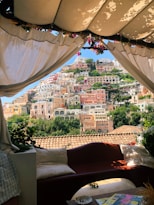 A picturesque view of a hillside covered in colorful buildings, seen through a shaded area with draped white curtains. The seating area in the foreground has a cozy, inviting atmosphere.