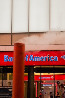 A Bank of America branch with a prominent red sign and blue lettering. A large steam pipe is emitting white steam in front of the building. There are people standing in front of the bank, visible through the windows.