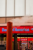 A Bank of America branch with a prominent red sign and blue lettering. A large steam pipe is emitting white steam in front of the building. There are people standing in front of the bank, visible through the windows.