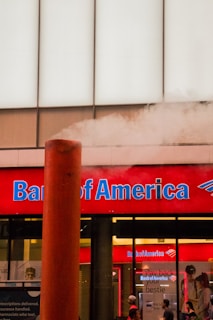 A Bank of America branch with a prominent red sign and blue lettering. A large steam pipe is emitting white steam in front of the building. There are people standing in front of the bank, visible through the windows.