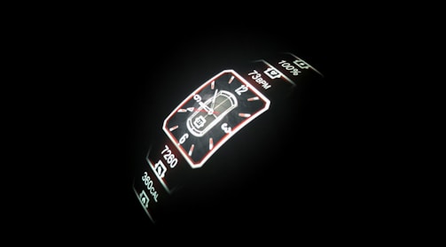 A digital smartwatch display is illuminated against a dark background. The screen features a digital clock with both analog and digital formats, including health metrics like step count, heart rate, and calorie count.
