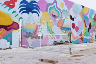 A vibrantly painted wall featuring abstract and geometric shapes in various colors, visible behind a chain-link fence topped with barbed wire. The ground is paved and slightly worn, and a 'Tow Away' sign is attached to the fence.