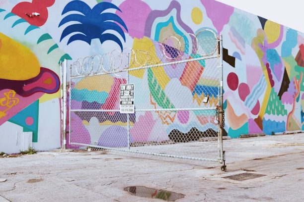 A vibrantly painted wall featuring abstract and geometric shapes in various colors, visible behind a chain-link fence topped with barbed wire. The ground is paved and slightly worn, and a 'Tow Away' sign is attached to the fence.