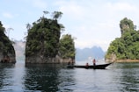 A picturesque scene of calm waters with several towering limestone cliffs covered in lush greenery. A traditional long-tail boat with people wearing life jackets is floating on the water in the foreground, creating a serene and tranquil atmosphere.