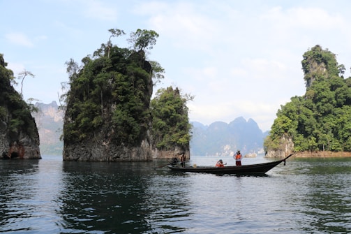 A picturesque scene of calm waters with several towering limestone cliffs covered in lush greenery. A traditional long-tail boat with people wearing life jackets is floating on the water in the foreground, creating a serene and tranquil atmosphere.
