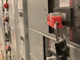 Close-up view of an electrical panel with a lockout tagout device attached. The panel is gray with multiple circuit breakers, and a red lock is secured with a Master Lock padlock for safety protocols.