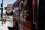 A large truck painted with a recognizable soda brand logo is parked on a city street. There are several cars driving on the road, and a rental bicycle is stationed nearby. The scene appears to be in an urban environment with tall buildings in the background and clear sky above.