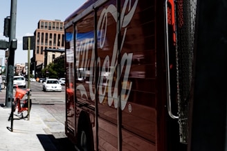 A large truck painted with a recognizable soda brand logo is parked on a city street. There are several cars driving on the road, and a rental bicycle is stationed nearby. The scene appears to be in an urban environment with tall buildings in the background and clear sky above.