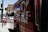 A large truck painted with a recognizable soda brand logo is parked on a city street. There are several cars driving on the road, and a rental bicycle is stationed nearby. The scene appears to be in an urban environment with tall buildings in the background and clear sky above.