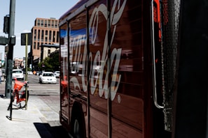 A large truck painted with a recognizable soda brand logo is parked on a city street. There are several cars driving on the road, and a rental bicycle is stationed nearby. The scene appears to be in an urban environment with tall buildings in the background and clear sky above.