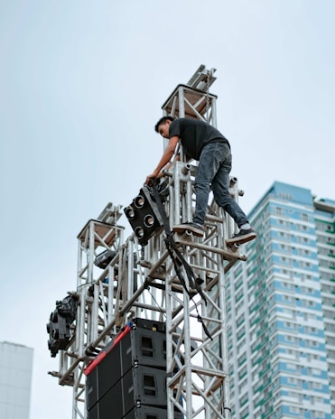 A person is climbing a metal scaffolding structure while setting up or adjusting equipment, likely for a stage or event. The background features a tall modern building with a clear sky above.