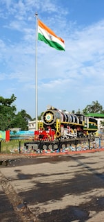 A vintage steam locomotive is displayed on a platform, surrounded by a decorative fence. Above it, a tall flagpole proudly flies the Indian national flag. The surrounding area includes some greenery and buildings under a clear blue sky with scattered clouds.