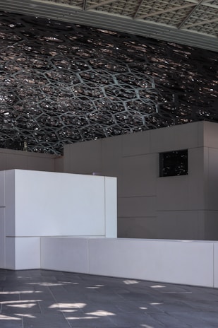 A geometric ceiling design featuring intricate latticework creates a pattern of shadows on the modern, minimalist interior architecture. The structure includes large, smooth white rectangular panels and a dark tiled floor that reflects the dappled sunlight from above.