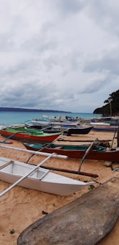 A sandy beach lined with several small traditional fishing boats, known as bangkas, facing a tranquil sea. The sky is overcast with gray clouds, suggesting an impending storm or recent rain, while the horizon features distant mountains. Some trees are visible near the shore.