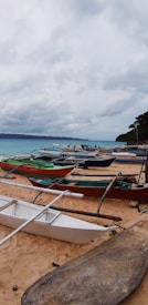 A sandy beach lined with several small traditional fishing boats, known as bangkas, facing a tranquil sea. The sky is overcast with gray clouds, suggesting an impending storm or recent rain, while the horizon features distant mountains. Some trees are visible near the shore.