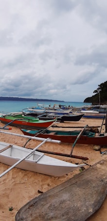 A sandy beach lined with several small traditional fishing boats, known as bangkas, facing a tranquil sea. The sky is overcast with gray clouds, suggesting an impending storm or recent rain, while the horizon features distant mountains. Some trees are visible near the shore.