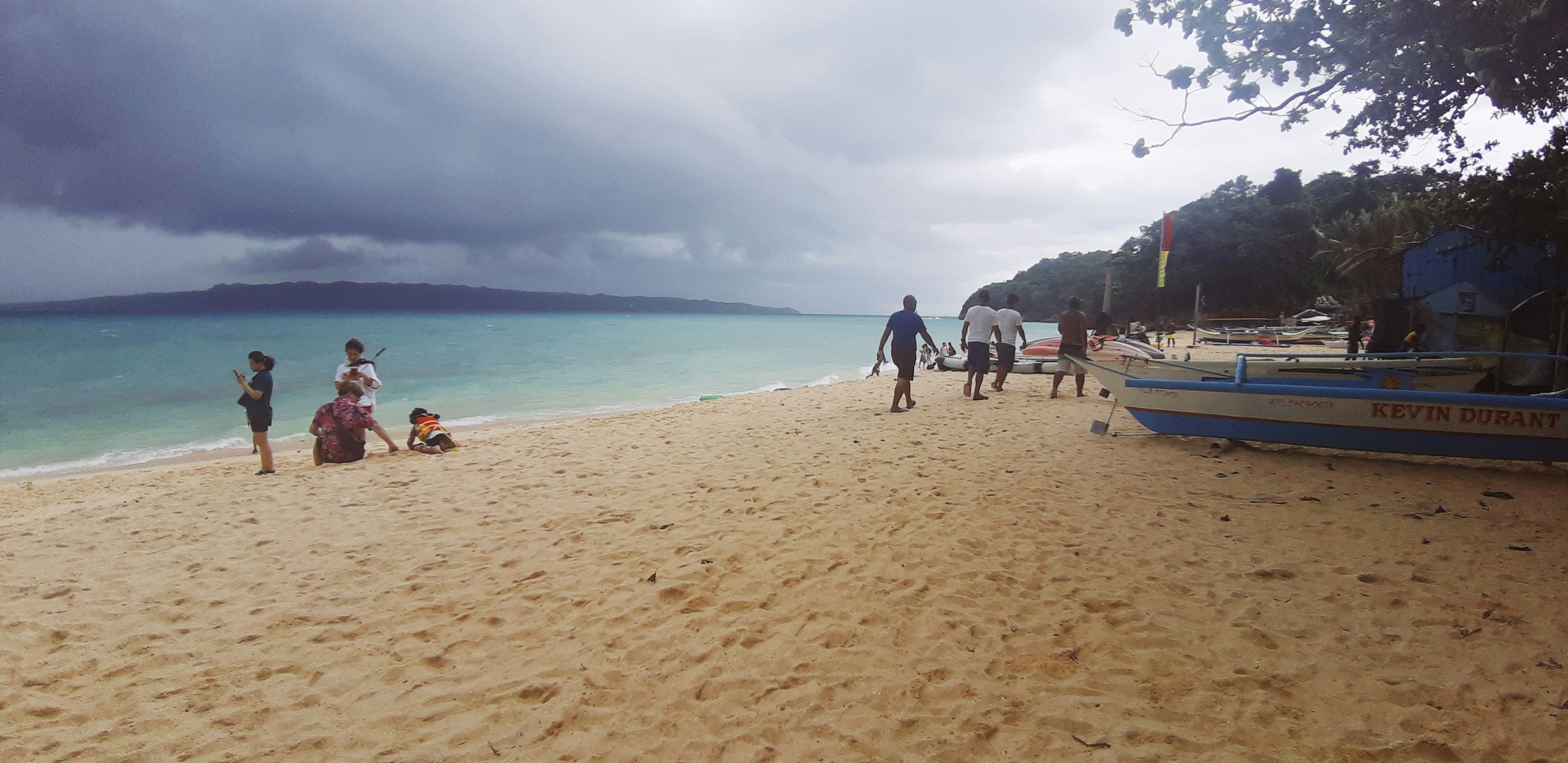 A group of people is walking and relaxing on a sandy beach. The sky is overcast with dark clouds, and the sea is a shade of turquoise. There are boats parked along the shore, with trees visible in the background.