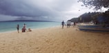 A group of people is walking and relaxing on a sandy beach. The sky is overcast with dark clouds, and the sea is a shade of turquoise. There are boats parked along the shore, with trees visible in the background.