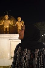 A person wearing a black hijab and patterned clothing stands in profile against a backdrop of golden statues of children holding hands around a globe. The scene is set at night with a dark sky, and there is a white pedestal supporting the statues.