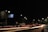 A nighttime urban street scene with bright streetlights illuminating the road, casting long light trails created by passing vehicles. A direction signpost indicates directions to Mulshi, Kolhapur, and Balewadi. The sky is dark, and some tree foliage is visible on the right.