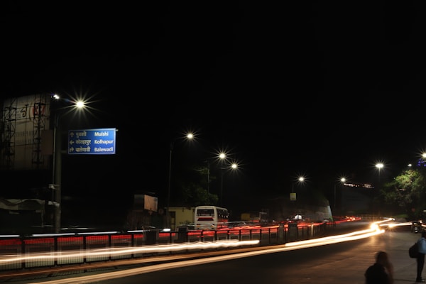 A nighttime urban street scene with bright streetlights illuminating the road, casting long light trails created by passing vehicles. A direction signpost indicates directions to Mulshi, Kolhapur, and Balewadi. The sky is dark, and some tree foliage is visible on the right.