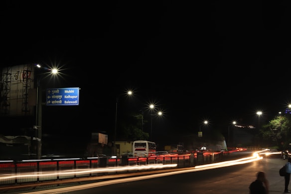 A nighttime urban street scene with bright streetlights illuminating the road, casting long light trails created by passing vehicles. A direction signpost indicates directions to Mulshi, Kolhapur, and Balewadi. The sky is dark, and some tree foliage is visible on the right.