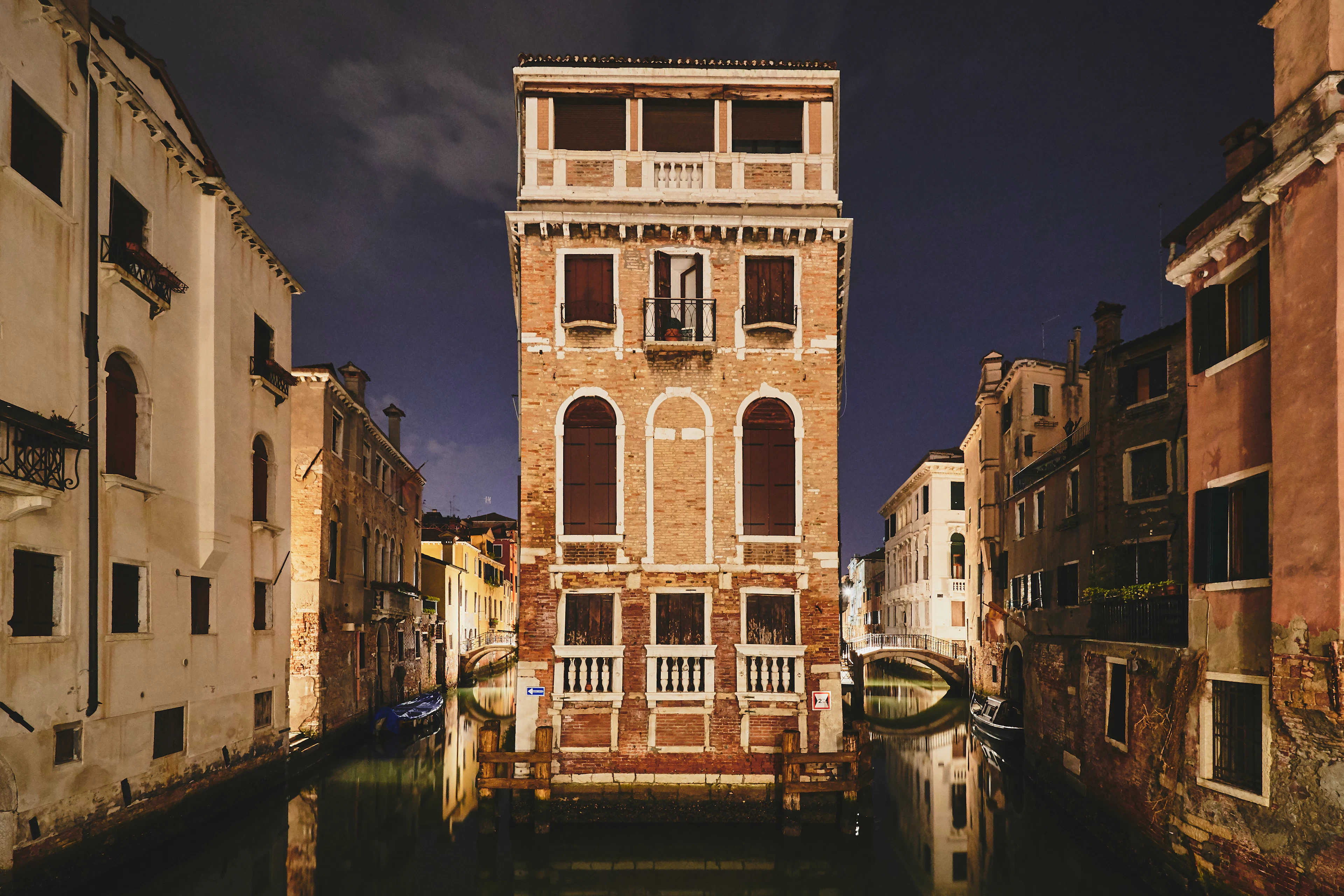 A brick palazzo glows under lamplight at night at the junction of two canals, its reflection rippling in the water.