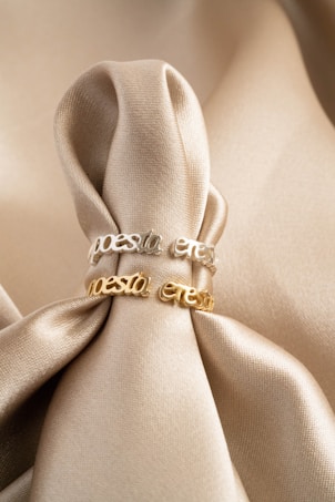 Two rings with the word 'pesta' are placed on a smooth, luxurious, beige satin fabric. The rings have two different finishes: one in silver and the other in gold. The fabric is elegantly gathered and draped around the rings, creating soft folds and a sophisticated backdrop.