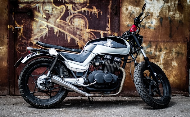 A vintage-style, black and white motorcycle is parked in front of a textured, rusty metal wall. The bike has a retro design with a prominent exhaust pipe and thick tires. Graffiti art is visible on the wall behind, adding an urban, gritty atmosphere.