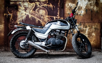 A vintage-style, black and white motorcycle is parked in front of a textured, rusty metal wall. The bike has a retro design with a prominent exhaust pipe and thick tires. Graffiti art is visible on the wall behind, adding an urban, gritty atmosphere.
