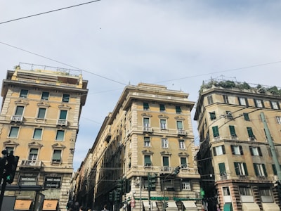 Three large, ornate residential buildings are viewed from street level. They have distinct stone facades, decorative moldings, and numerous windows with green shutters. Rooftop gardens are visible on the top floors. Trolley cables stretch overhead against a pale blue sky.