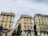 Three large, ornate residential buildings are viewed from street level. They have distinct stone facades, decorative moldings, and numerous windows with green shutters. Rooftop gardens are visible on the top floors. Trolley cables stretch overhead against a pale blue sky.