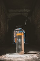 A cylindrical metal bollard stands in the center of a dark, narrow alleyway. Sunlight illuminates part of the bollard while casting shadows in the surrounding area. Beyond the alley, colorful facades of urban buildings are visible, featuring vibrant blue and orange tones.