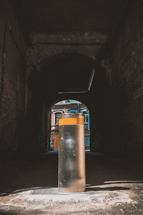 A cylindrical metal bollard stands in the center of a dark, narrow alleyway. Sunlight illuminates part of the bollard while casting shadows in the surrounding area. Beyond the alley, colorful facades of urban buildings are visible, featuring vibrant blue and orange tones.