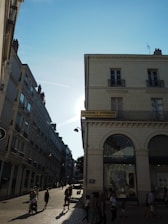 A sunlit street with people walking along a cobblestone path. On the right, there is a building with a sign that reads 'Cabinet Piroud' and has arched windows. The sky is clear with a visible contrail and the sunlight creates strong contrasts and shadows.